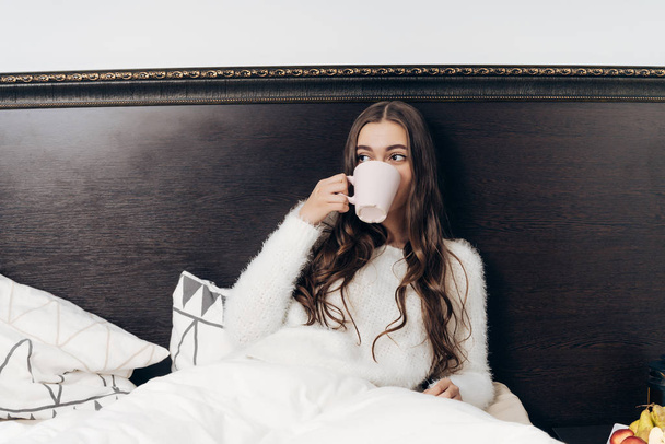 long-haired young girl sitting in bed early in the morning, having breakfast and drinking fragrant coffee - Фото, изображение