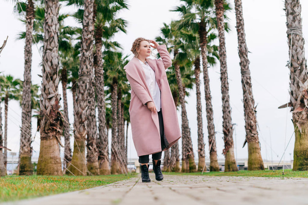 beautiful young girl in a pink fashion coat walks in the park against the backdrop of palm trees, enjoys the warm spring weather - Фото, изображение