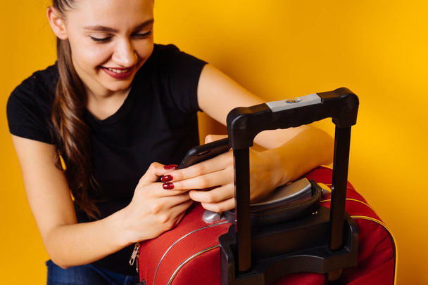 menina feliz vai em uma viagem, com uma grande mala vermelha, olha para o smartphone e sorri
 - Foto, Imagem