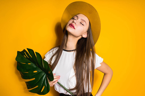 confident long-haired girl model in a fashionable hat posing on a yellow background, holding a green leaf, closing her eyes - 写真・画像