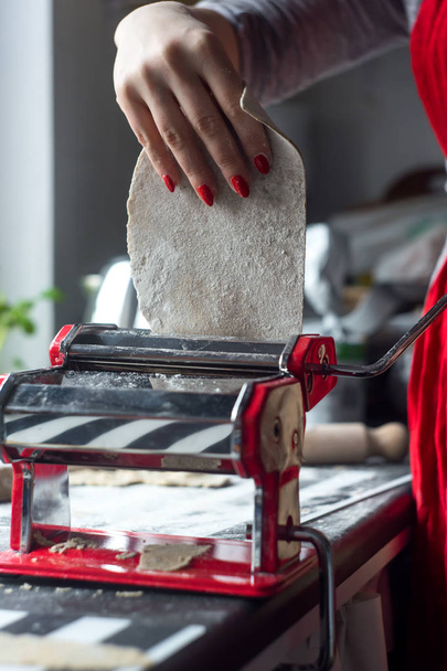 Frau rollt Teig für Pasta am Küchentisch mit rotem Nudelmahlwerk. - Foto, Bild