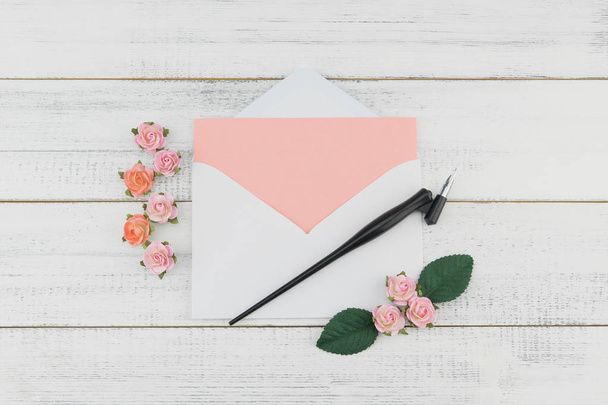 Чистая розовая открытка в белом конверте и косой ручкой украсить розовой розовой бумаги цветы на белом фоне дерева
 - Фото, изображение