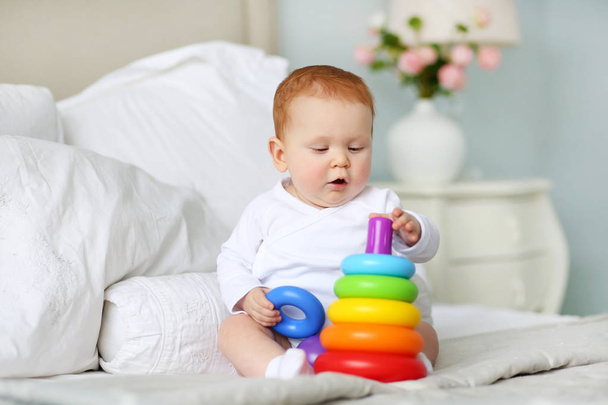 Cute baby playing with colorful rainbow toy pyramid sitting on bed in white sunny bedroom. Toys for little kids.  Child with educational toy. Early development. - Foto, immagini