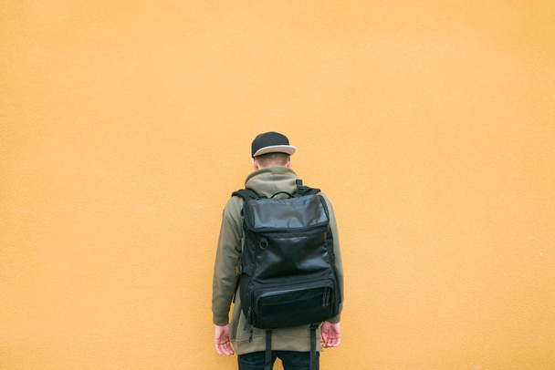 Um homem elegante nas costas com uma mochila no fundo de uma parede laranja. Um jovem em um camarim e uma grande mochila fica no fundo da parede
 - Foto, Imagem