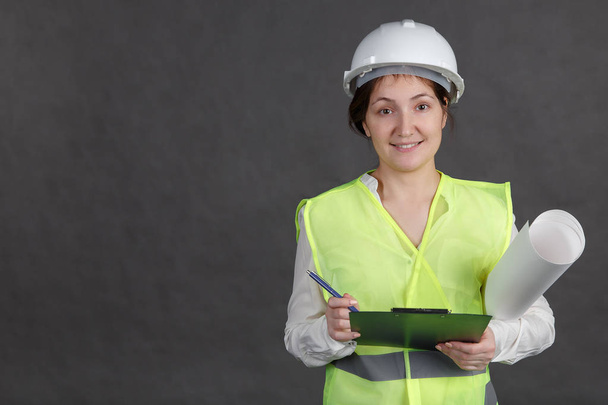 Young woman engineer in protective helmet and vest with projects. Makes notes in the tablet - Photo, Image
