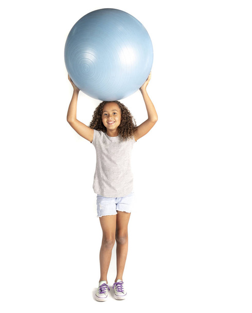 Jovem criança brincalhão ou pré-adolescente menina birracial com cabelo encaracolado marrom jogando ou se exercitando com uma grande bola de borracha segurando-o sobre a cabeça e rindo
.  - Foto, Imagem