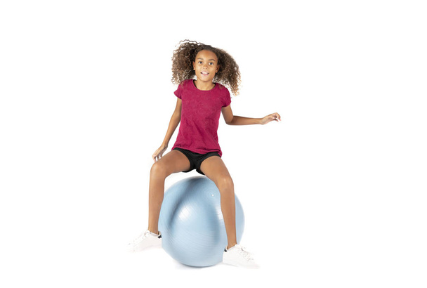 Niño juguetón o niña pre adolescente birracial con el pelo rizado marrón jugando o haciendo ejercicio en una pelota de goma grande
 - Foto, Imagen