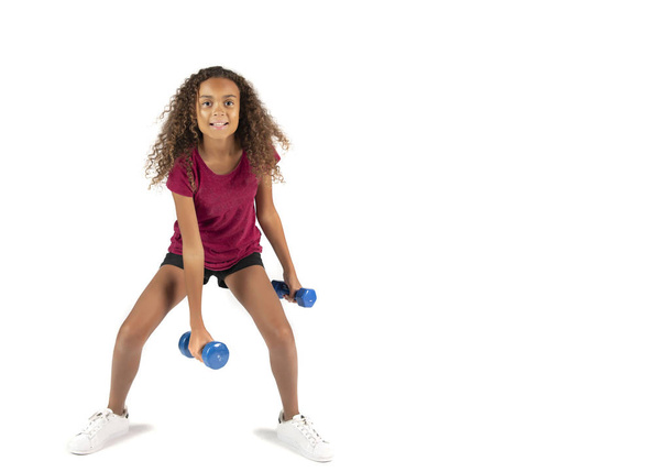 Молодой игривый ребенок или доподростковая расовая девочка с каштановыми кудрявыми волосами тренирующаяся с маленькими голубыми гантелями
 - Фото, изображение