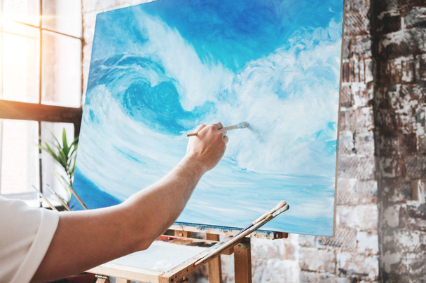 Male hand of artist closeup holding paintbrush on background of canvas on easel. Painter drawing ocean wave in studio. Flare effect - Фото, изображение