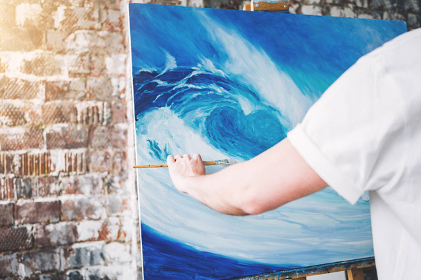Male hand of artist holding paintbrush on background of canvas on easel. Professional painter drawing ocean wave in loft studio. Flare effect - Фото, зображення