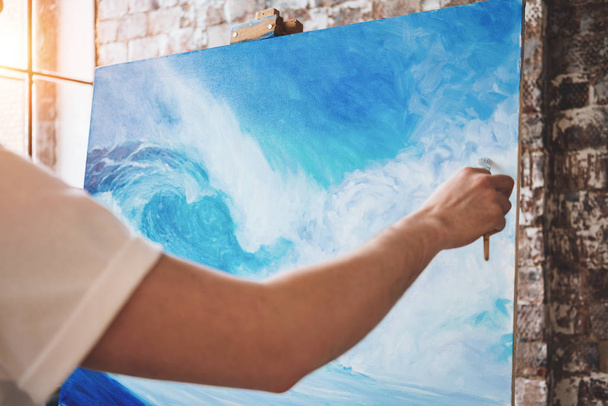 Male hand of artist closeup holding paintbrush on background of canvas on easel. Professional painter drawing ocean wave in loft studio. Flare effect - Фото, изображение