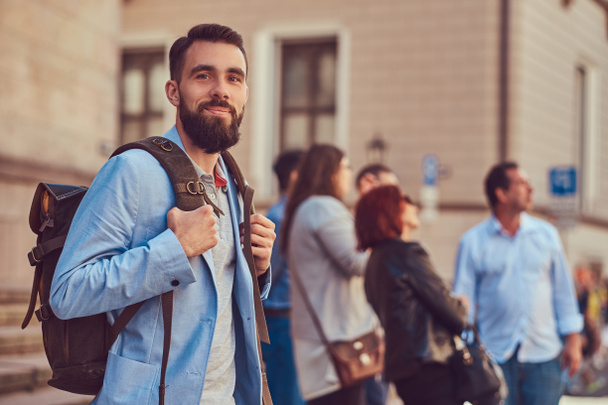 Турист с бородой и прической, в повседневной одежде, держит рюкзак во время экскурсии по Европе
. - Фото, изображение