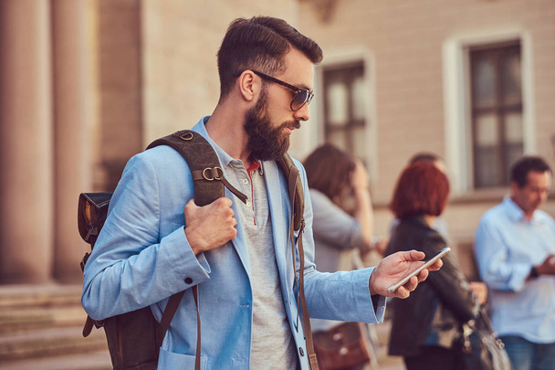 Un turista con barba completa y corte de pelo, con ropa casual y gafas de sol, sostiene una mochila y mensajes de texto en un teléfono inteligente, de pie en una calle antigua, durante la excursión en Europa
. - Foto, Imagen