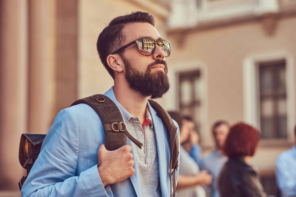 A tourist with a full beard and haircut, wearing casual clothes and sunglasses, holds a backpack, standing on an antique street, during the excursion in Europe. - Photo, image