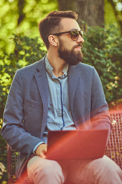 Ritratto ravvicinato di uno scrittore con la barba piena ed elegante taglio di capelli, che lavora con un computer portatile mentre è seduto su una panchina in un parco
. - Foto, immagini