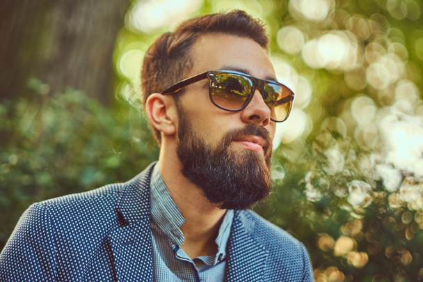 Retrato de cerca de un hombre barbudo con un corte de pelo elegante, con una chaqueta y gafas de sol, sentado en un banco en un parque de la ciudad
. - Foto, imagen