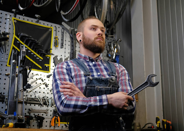 Portrait of a handsome stylish male with beard and haircut wearing a flannel shirt and jeans coverall, holds steel wrench, standing in a workshop against wall tools. - Foto, Imagen