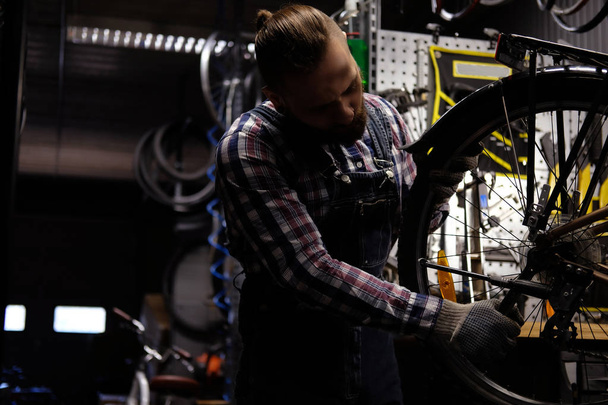 ein hübscher, stylischer Mann in Flanellhemd und Jeans-Overall, der in einer Werkstatt mit einem Fahrradrad arbeitet. Arbeiter montiert mit einem Schraubenschlüssel das Rad an einem Fahrrad in einer Werkstatt. - Foto, Bild