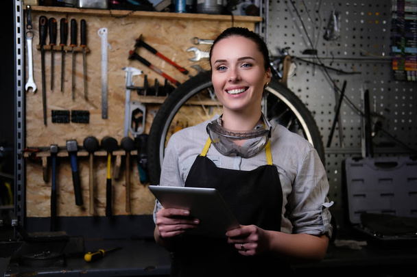 Portrait of a smiling beautiful brunette female wearing working clothes, apron and goggles, holds a tablet computer in a workshop. - Photo, image