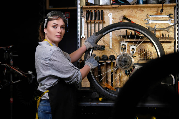 Beautiful female in working clothes, apron and goggles, repairs a bicycle wheel in a workshop. - Photo, Image