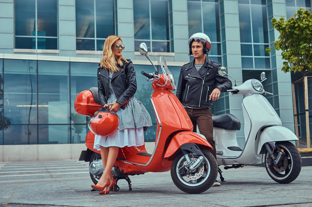 An attractive romantic couple, a handsome man and sexy female, standing with two retro Italian scooters against a skyscraper. - Photo, Image