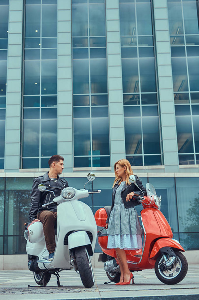 Attractive romantic couple, a handsome man and sexy female spend time together outdoors against a skyscraper. Walking with scooters during a date. - Photo, image