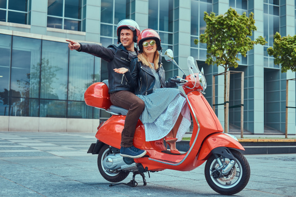 An attractive happy couple, a handsome man and sexy female riding together on a red retro scooter in a city. - Photo, Image