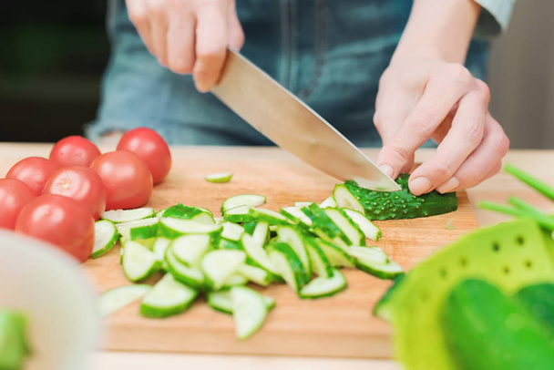close-up of female hands cut into fresh cut cucumbers on a wooden cutting board next to pink tomatoes. The concept of homemade vegetarian cuisine and healthy eating and lifestyle - 写真・画像