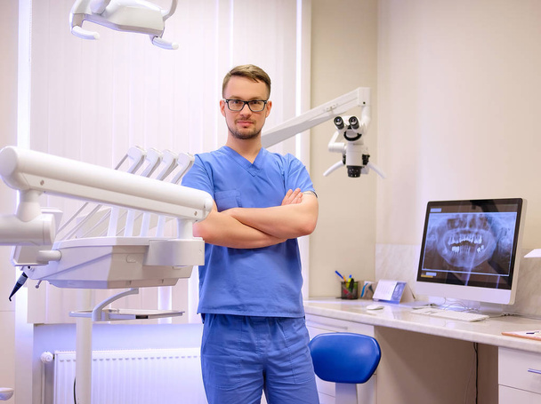 Portrait of a handsome dentist wearing a blue uniform, standing in a dentist clinic. Looking at camera. - Foto, Bild
