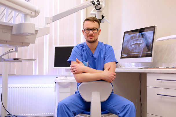 Portrait of a handsome bearded dentist male in glasses wearing a blue uniform, sitting in a dentist office. Looking at camera. - Foto, Bild