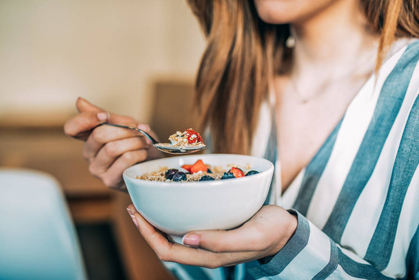Καλλιέργεια γυναίκα κοντά επάνω τρώγοντας μπολ βρώμης και τα φρούτα για πρωινό - Φωτογραφία, εικόνα