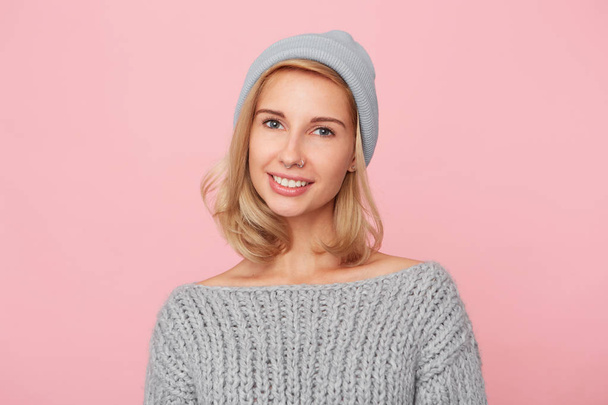 Portrait of young blonde woman, wears sweater and cap looks directly into camera and smiles. Shows her white teeth. Isolated over pink background - Foto, Imagen
