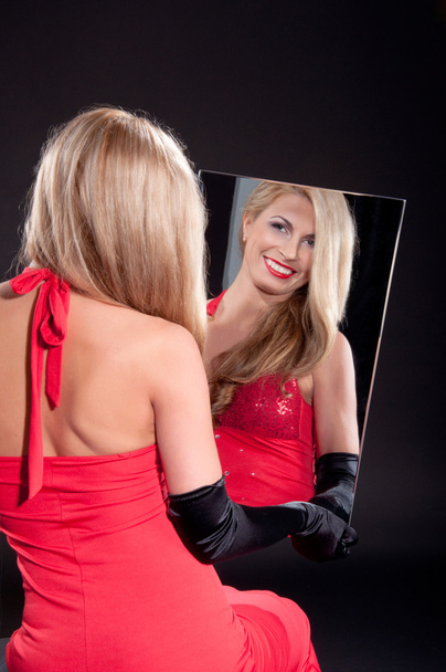 Beauiful young woman in red dress looks into the mirror on dark background - Фото, изображение