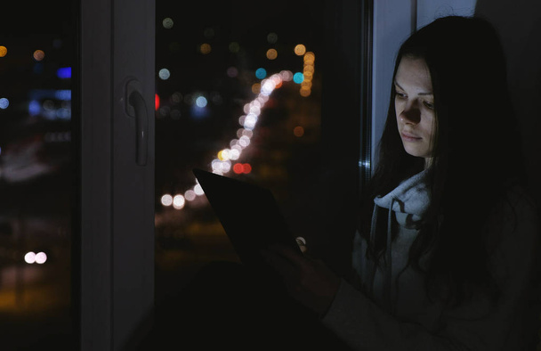 Woman sitting on the windowsill in the dark night and using tablet for web. - Foto, Bild