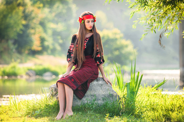 girl in a designer embroidered suit with patterns sits on the stone near the river - Photo, Image