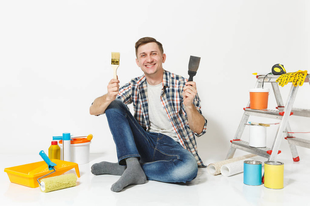 Happy smiling male sitting on floor with brush, putty knife instruments for renovation apartment room isolated on white background. Wallpaper, gluing accessories, painting tools. Repair home concept. - Photo, Image