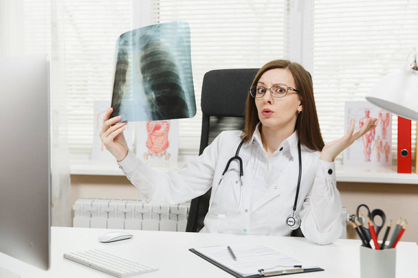 Ärztin sitzt am Schreibtisch, arbeitet am Computer, hält Röntgenbilder der Lungen, Fluorographie, Röntgen im Lichtbüro im Krankenhaus. Frau im Arztkittel, Stethoskop im Sprechzimmer. Medizinkonzept. - Foto, Bild