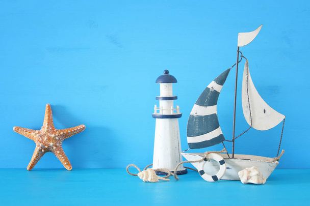 nautical concept with white decorative sail boat, lighthouse, starfish, seashells over blue wooden table and background. - Photo, Image
