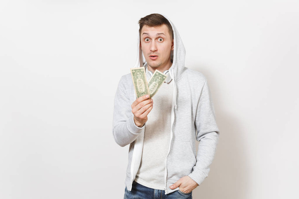 Young handsome man student in t-shirt, blue jeans and light sweatshirt with hood with headphones holds two one-dollar bills and surprised looks isolated on white background. Concept of emotions - Photo, Image