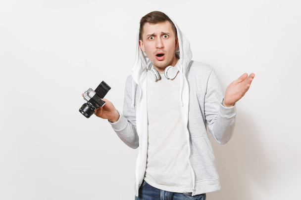 Jonge knappe verrast mannelijke student in t-shirt en lichte sweatshirt met capuchon met koptelefoon houdt retro camera en verspreidt zijn handen geïsoleerd op een witte achtergrond. Concept van fotografie, hobby - Foto, afbeelding