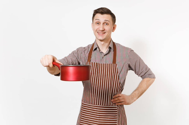 Jong glimlachen man chef-kok of kelner in gestreepte bruine schort, shirt bedrijf rode lege stewpan, pan of pot geïsoleerd op een witte achtergrond. Mannelijke huishoudster of houseworker. Keukengerei en keuken concept. - Foto, afbeelding