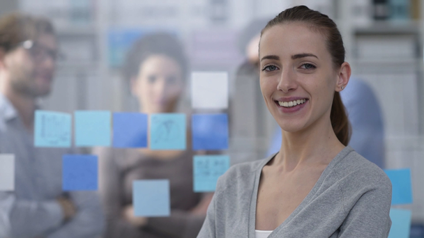 Joven mujer sonriente posando en la oficina y el equipo de negocios trabajando en el fondo
 - Metraje, vídeo