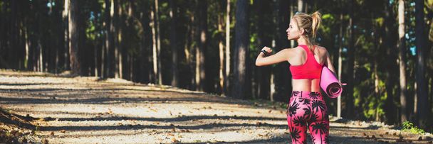 Young fit athletic woman in a forest wearing smart watch and holding yoga mat, walking away from the camera. Mind and body web banner. - Foto, imagen