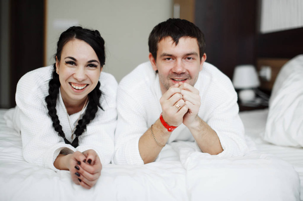 Nuori aviomies ja vaimo valkoiset takit leikkiä ja hymyillä sängyssä hotellihuoneessa. Sulje se.
. - Valokuva, kuva