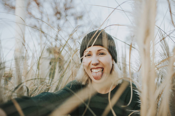Portrait of cute blonde woman with a knit hat on taking out the tongue, surrounded of reed plants in nature. - Foto, Bild