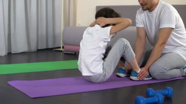 Boy doing sit-ups at home with help of father, family training, sport childhood - Footage, Video