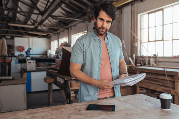 Portrait of skilled young woodworker standing at workbench in his carpentry studio using digital tablet and holding notebook of designs - Foto, Imagem