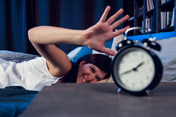 Фото недовольной женщины с бессонницей, протягивающей руку к будильнику ночью
 - Фото, изображение