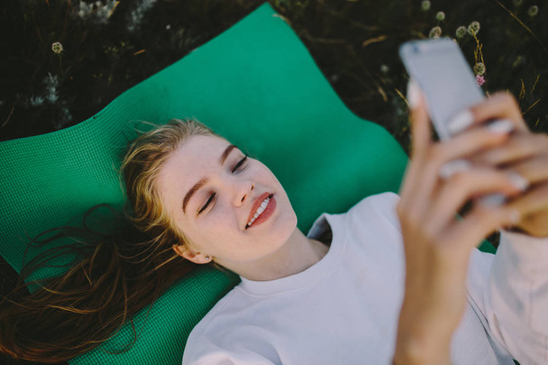 young beautiful blond girl teenager lying on green yoga mat with phone. Smiling girl used smartphone outdoor. Closeup - Фото, зображення