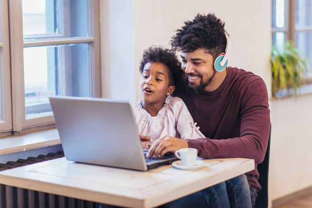 Молодий чорний батько сидить разом зі своєю дочкою, вони використовують ноутбук
 - Фото, зображення